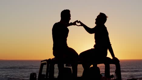 happy caucasian gay male couple sitting on car making heart shape with hands at sunset on the beach