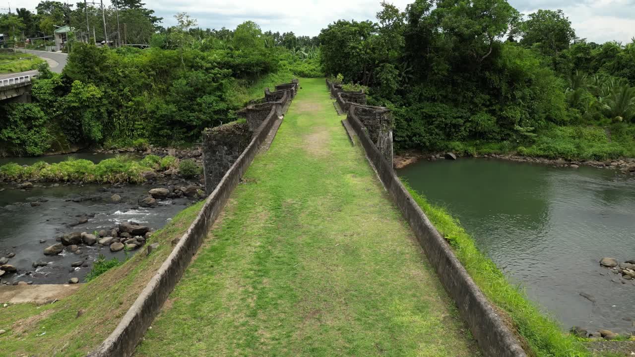 A fast push in drone shot over the historic Puente de Malagonlong Bridge's mossy walk way, spanning a rocky river amid lush tropical greenery of Tayabas, Quezon Province Philippines