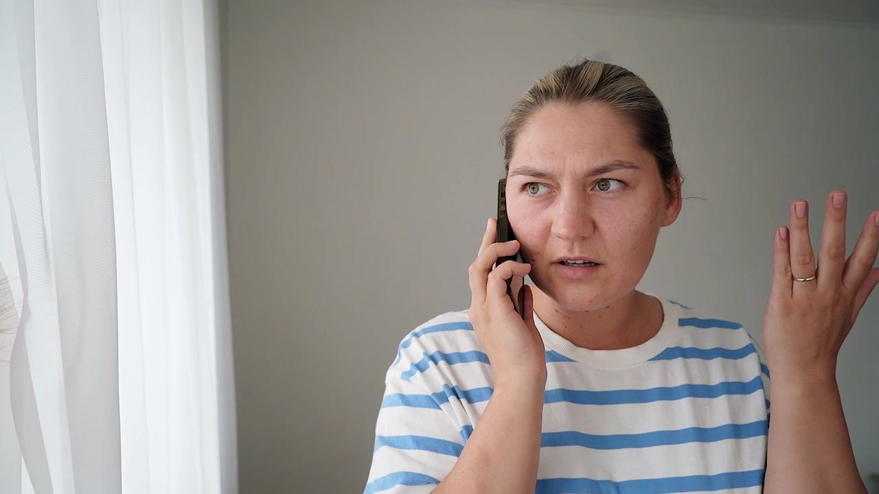 Irritated customer in a striped T-shirt talks on a smartphone indoors, reacting with frustration during a tense call.