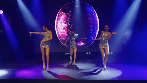 three female singers performing on a stage with a disco ball and bright lights