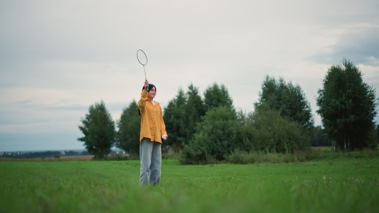 woman in yellow jacket moving in meadow, tossing small objects and reaching with controlled gestures slow fluid training sequence across green field under overcast sky, distant trees framing scene