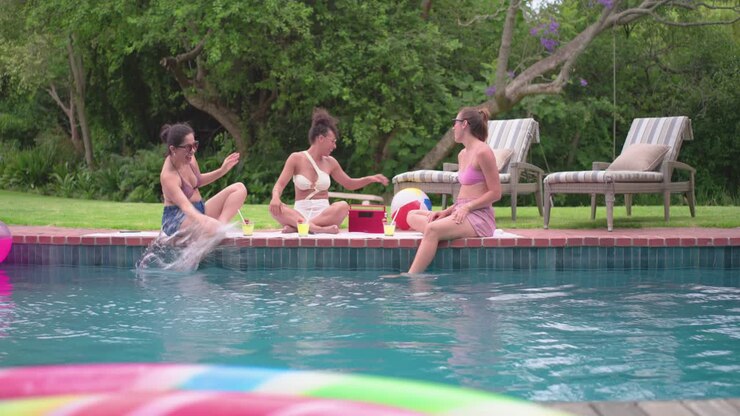 Diverse female friends relaxing at pool in bikinis and shades nudging ball scooting causing splash
