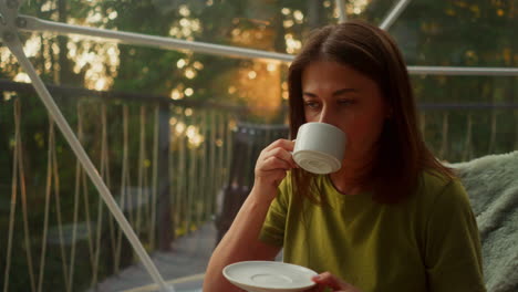 mujer tranquila bebiendo café disfrutando de la vista desde la ventana panorámica al atardecer en una noche cálida. vacaciones tropicales en un edificio turístico moderno rodeado de árboles de diferentes tipos