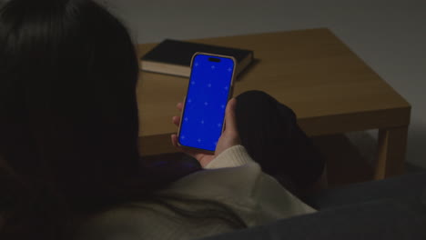 Over The Shoulder Shot Of Woman At Home Sitting On Sofa With Blue Screen Mobile Phone Scrolling Through Internet Or Social Media