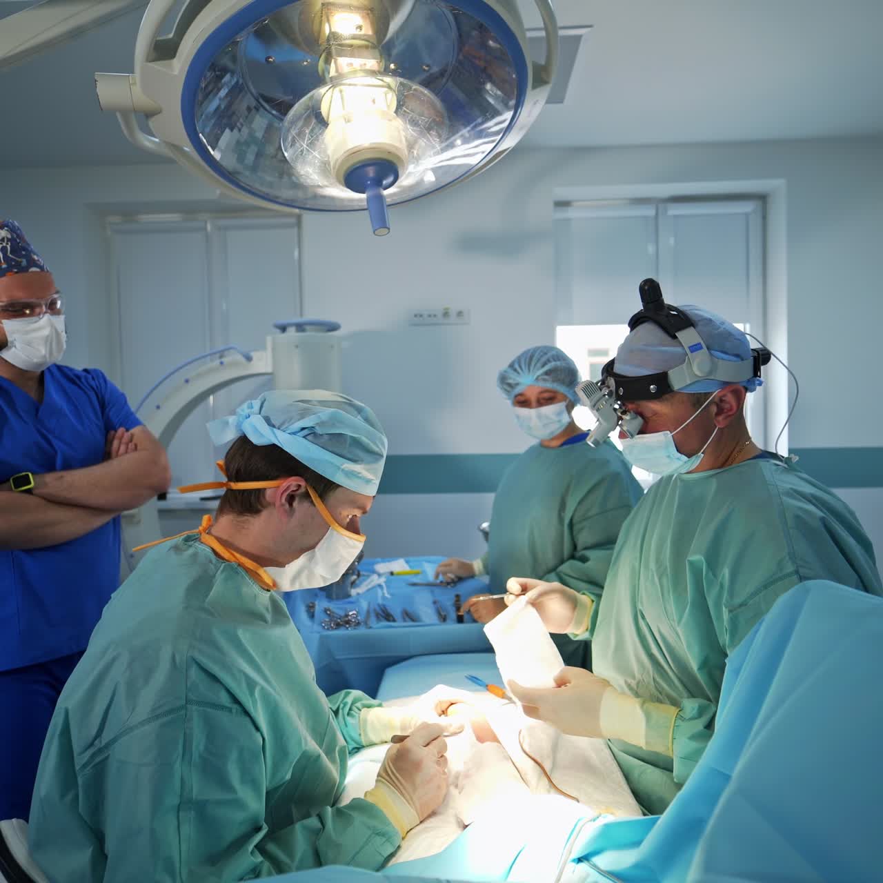 Collaboration of two surgery professionals at operation. Doctors sit bending over the patient at operational table. Anesthesiologist standing beside watching the process
