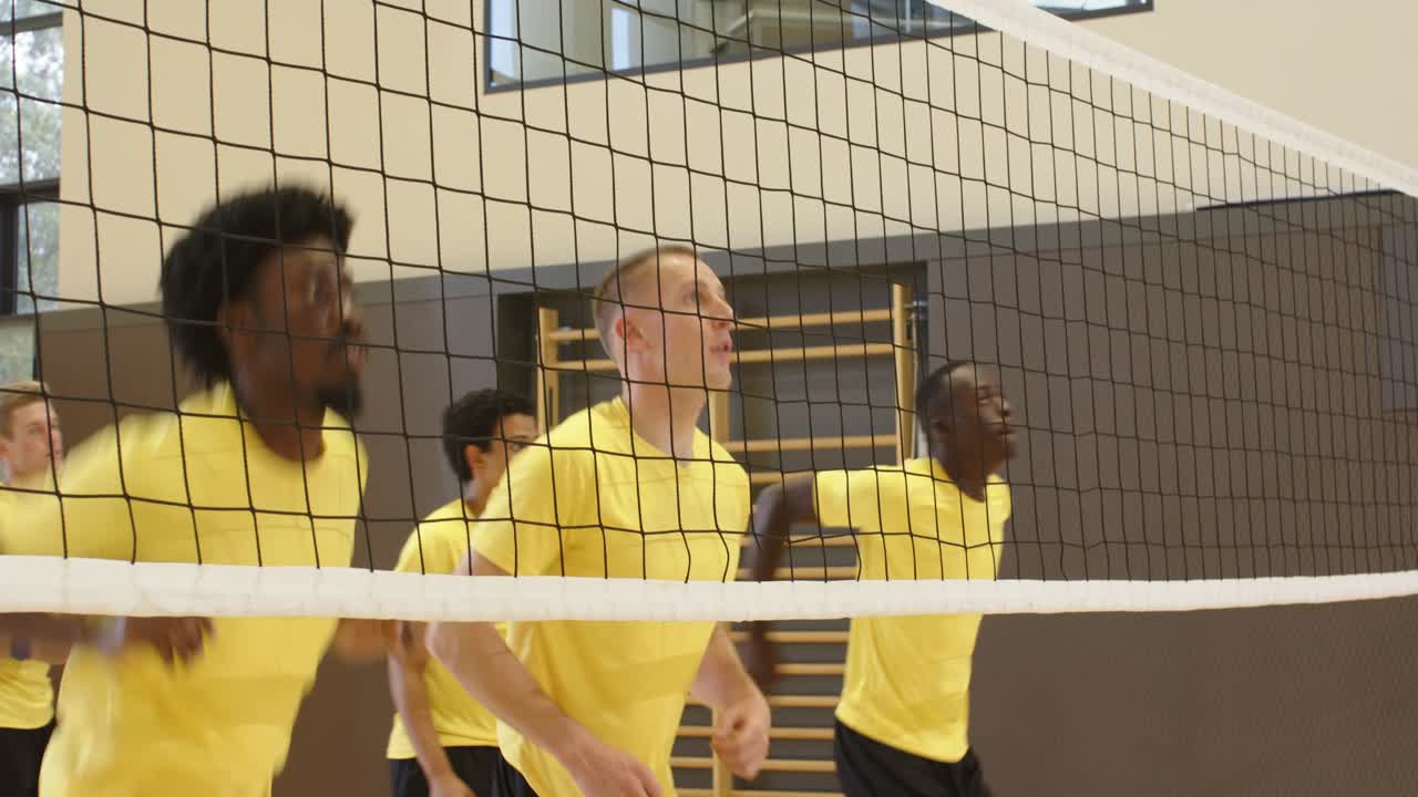 Seks mandlige spillere i gule trøjer hopper og blokerer med volleyball ved nettet, mens træningsøvelsen starter i idrætshallen.