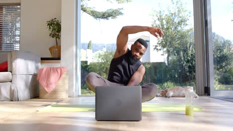 hombre afroamericano enfocado practicando yoga usando una computadora portátil en casa soleada, cámara lenta