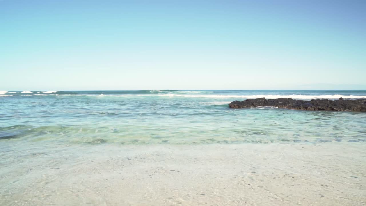 작은 파도가 노란 모래 해변에서 무릎을 꿇는 동안 용암 선반 암초 형성 위로 굴러가는 큰 흰색 모자를 씌운 파도