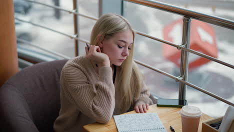 joven sentada escribiendo pensativamente con la mano en la mandíbula, involucrada en un pensamiento profundo, con una taza de café en la mesa y un fondo que muestra un entorno urbano ocupado en invierno