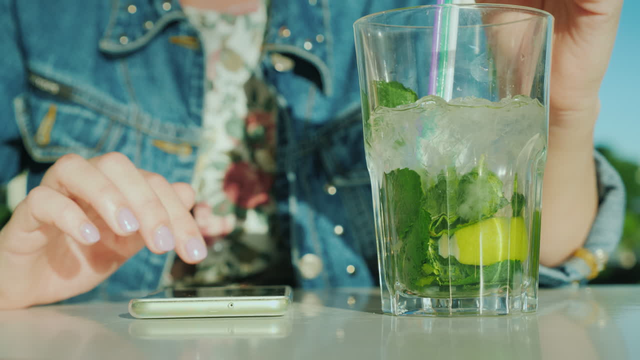 una mujer usa un teléfono inteligente a continuación hay un vaso con un cóctel fresco en el marco solo se ven las manos