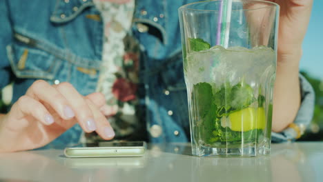 a woman uses a smartphone next is a glass with a cool cocktail in the frame are visible only hands