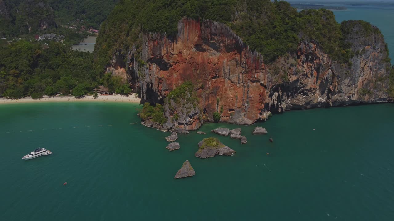 Aerial shot of Ao Nang Beach and its dramatic red limestone cliffs rising above the turquoise sea, with boats and a yacht anchored near the tropical shoreline in Krabi, Thailand.