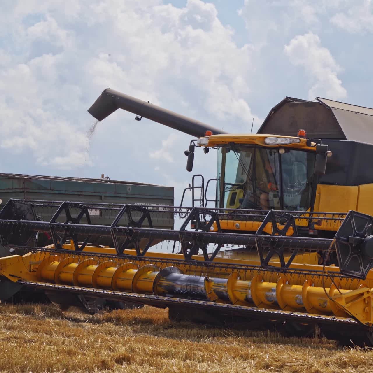 Modern combine pouring out crop into cart on the field. Yellow combine harvester in action during seasonal works under blue sky.