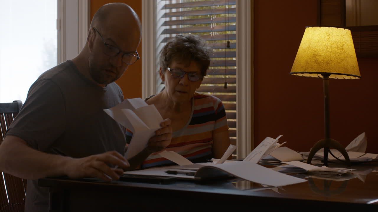 Adult son and his senior mother sit together at a dining table in a warmly lit home, sorting through papers and bills. Caregiver son helps mother count, and calculate receipts under table lamp light.