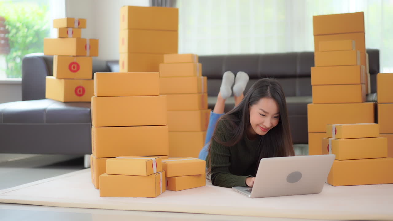 Asian woman on floor with laptop computer surrounded by boxes. Young lady filling online orders for internet based business. Packages ready to be shipped or just received by girl shopping
