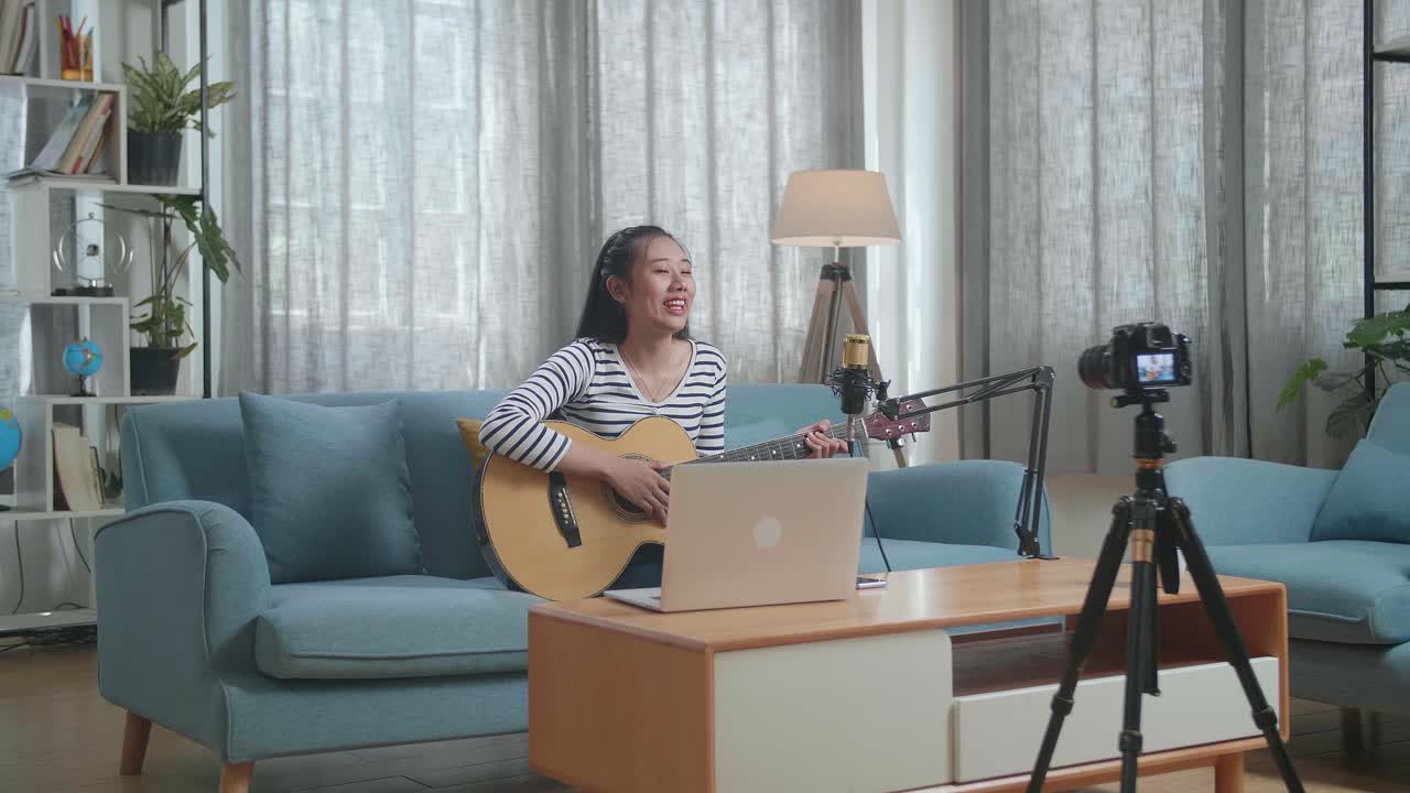 아시아 여성이 노트북을 들고 손을 흔들며 카메라에 대화하면서 라이브 스트리밍을 하며 집에서 기타를 연주하고 있습니다.