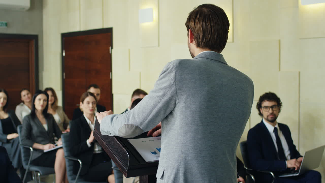 Rear view of Caucasian businessman speaker on a podium talking in a conference room in front to many people