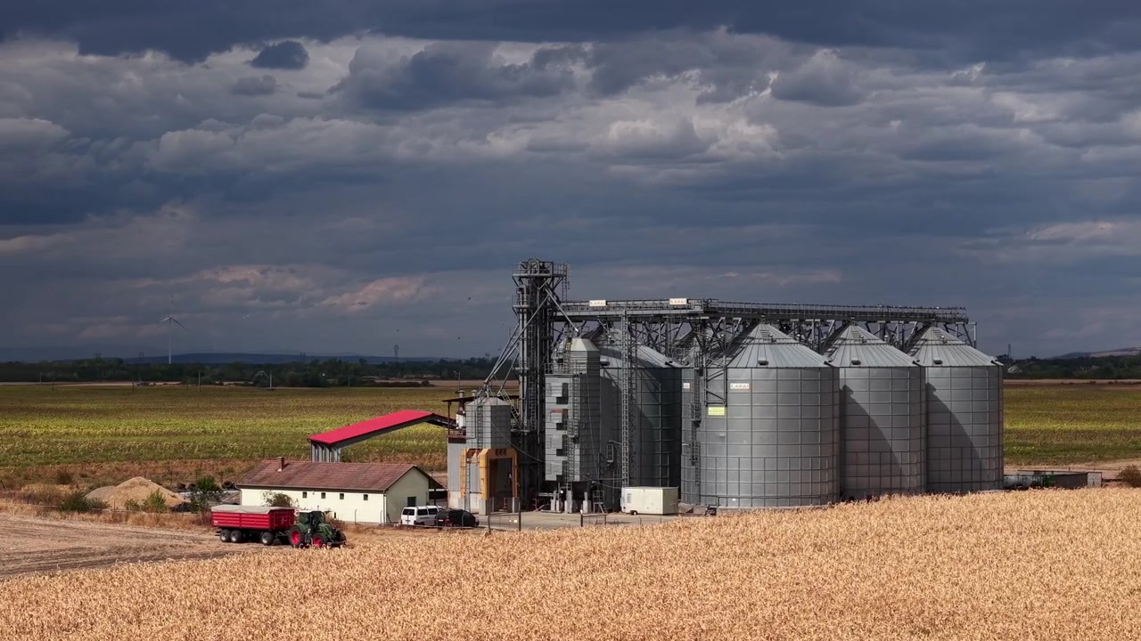 Uma grande instalação industrial de armazenamento de grãos domina uma vasta paisagem agrícola sob um céu nublado e sombrio com nuvens de tempestade.