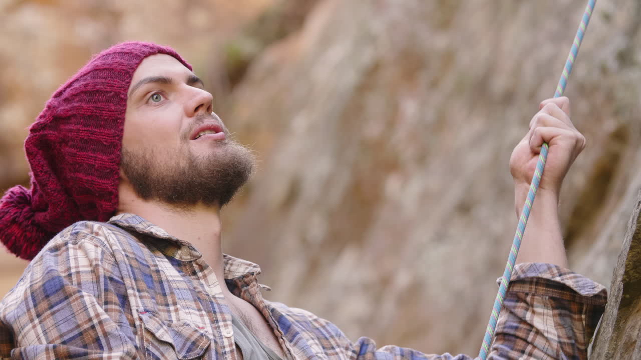 Medium close up of bearded male mountaineer wearing flannel shirt and red beanie belaying a rope while looking up and talking to his rock climbing partner in realtime