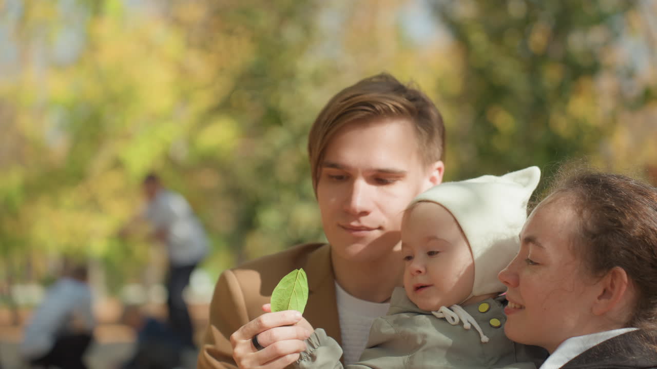 Herbstweg, kaukasisches Ehepaar bietet Baby ein Blatt an. Vater hält Baby, während Mutter lacht, leuchtendes Laub und warmes Sonnenlicht, spielerischer Austausch, freudige Familienbindung, authentisches Porträt.