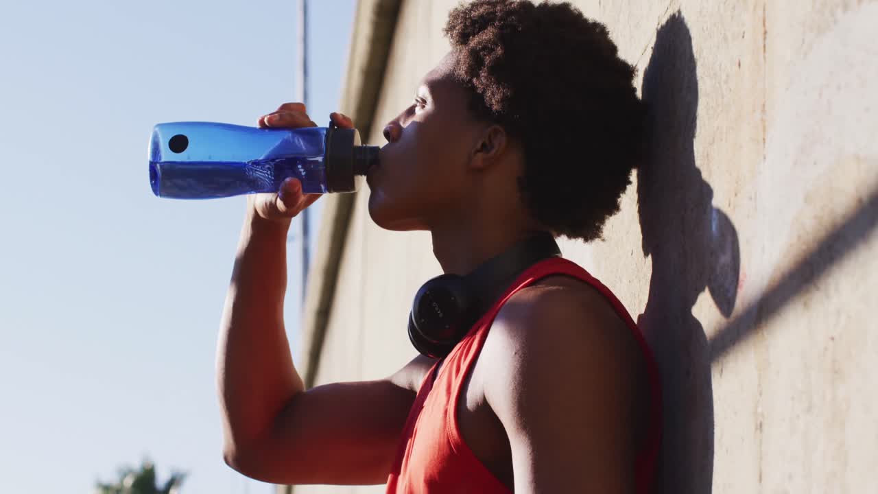 hombre afroamericano en forma haciendo ejercicio en la ciudad tomando un descanso, apoyado en la pared bebiendo agua