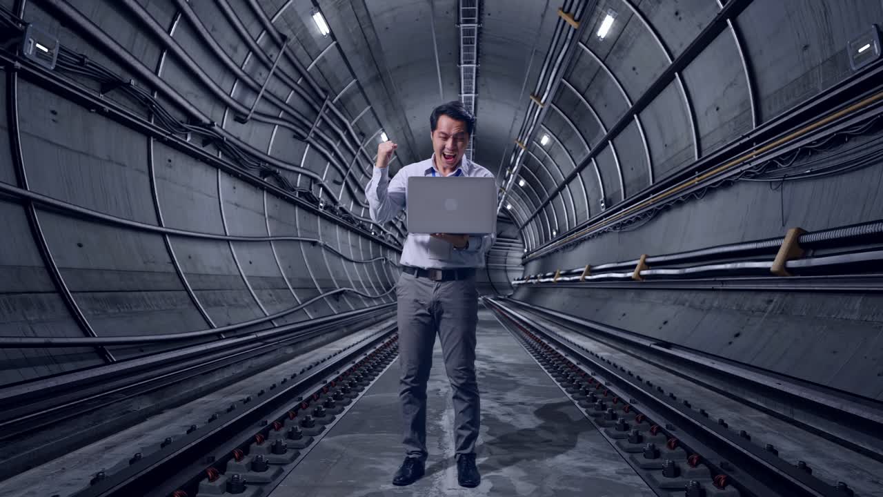 Full Body Of An Asian Male Professional Worker Standing With His Laptop In Underground Subway Tunnel, He Raises His Fist Up With Screaming Goal