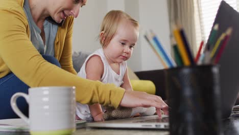caucasian mother holding her baby using laptop and talking on smartphone while working from home