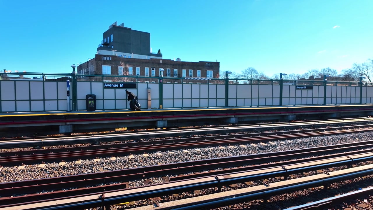 Avenue M metroplatform og spor i Brooklyn. Bred dagsudsigt viser flere skinner, stationsskilte og en rejsende, der går under en klar blå vinterhimmel