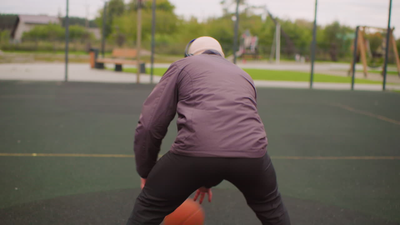 Older man dribbling basketball outdoors wearing jacket and cap with headphones, practicing low dribble and footwork on empty suburban court, focused posture and steady rhythm, hoop visible in distance