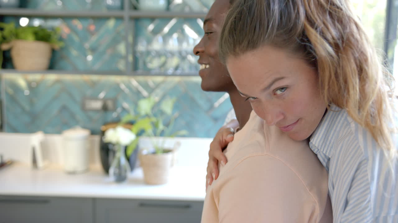 diverse couple embracing in kitchen, woman smiling and looking at camera, copy space