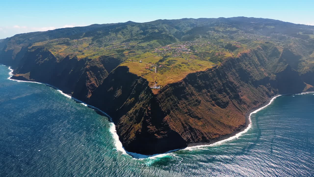 Triangular high rock deeps into the blue waterscape of the Atlantic Ocean. A town is located on the top of the mountain. Top view on the Madeira Islands, Portugal.