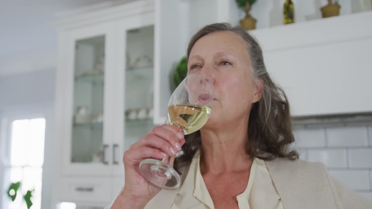 Senior caucasian woman standing in kitchen enjoying drinking a glass of white wine