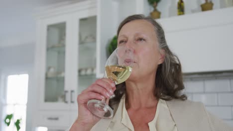 Senior-caucasian-woman-standing-in-kitchen-enjoying-drinking-a-glass-of-white-wine