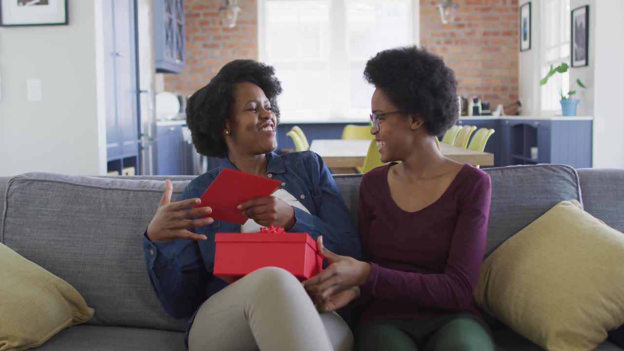 Madre e figlia afroamericane felici sedute sul divano, che si scambiano un regalo.