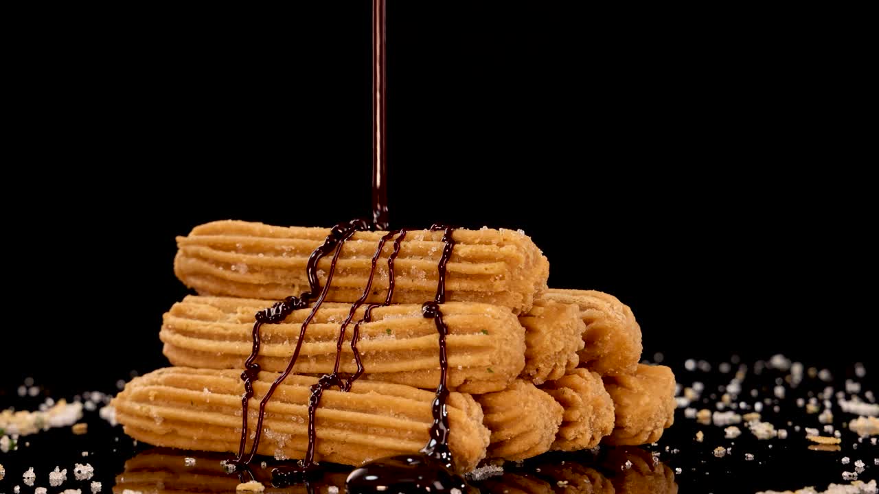 Golden churros are stacked on a reflective surface as rich chocolate sauce is poured over them, with dramatic lighting and a dark backdrop