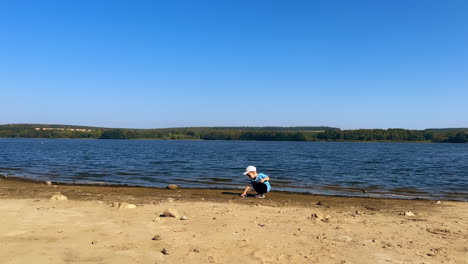 Lovely Caucasian toddler spending time on the river bank in summer. Kid picks up the stone to throw it into water.