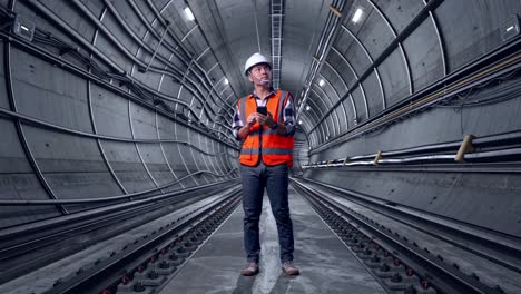 full body of asian male engineer with safety helmet using smartphone and looking around while standing in underground subway tunnel