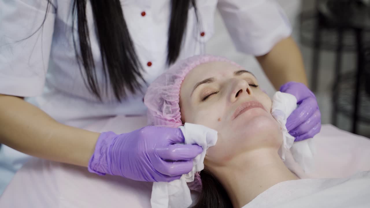A beautician in gloves holds sanitary napkins in her both hands and cleans the skin of the client's face with it before the procedure. Close-up.