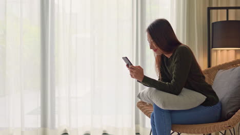 Young-woman-using-a-mobile-phone-and-typing-a-message-or-surfing-the-internet-while-sitting-on-a-chair-in-her-living-room