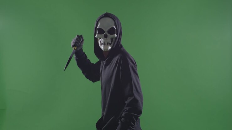 страшный мужчина в пуловерке с капюшоном, носящий маску хэллоуина, пронзает ножом и поворачивает к камере, делая угрожающий жест на зеленом фоне экрана.