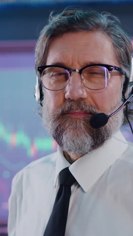 Bearded-middle-aged-man-with-headset-and-glasses-looking-at-camera-while-working-in-stock-exchange-office.-Vertical-shot