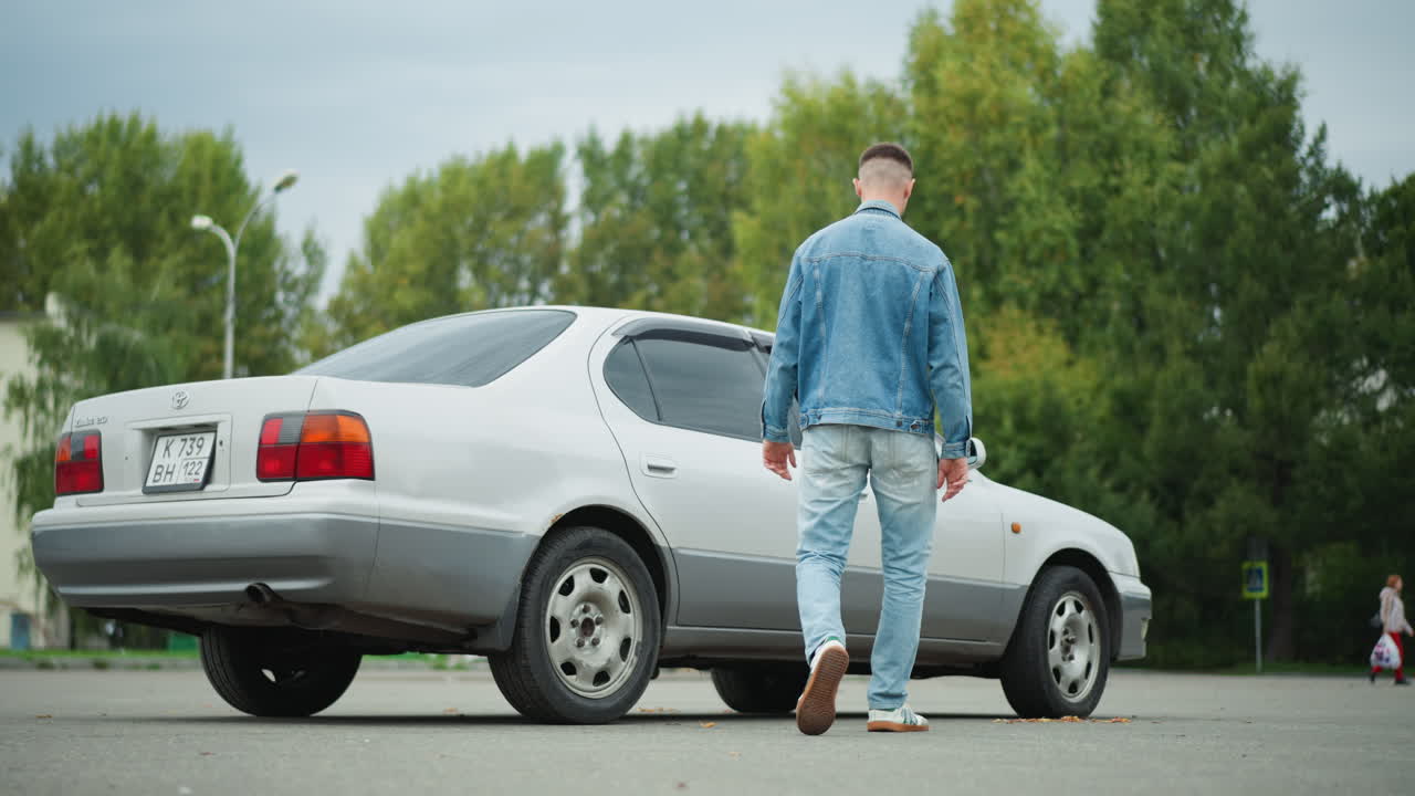 Un giovane uomo torna alla sua berlina color argento e si prepara a guidare. Indossa una giacca di jeans e jeans, ha un atteggiamento sicuro mentre chiude la portiera e regola lo specchietto retrovisore. Ampio parcheggio con alberi verdi e condomini