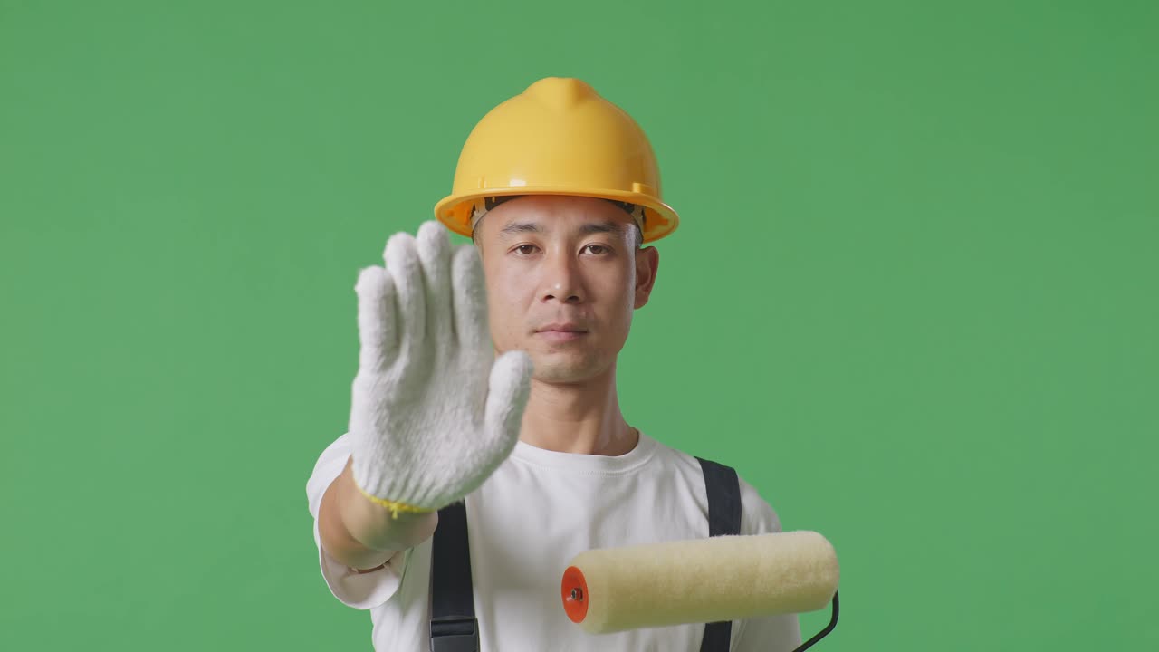 Close Up Of Asian Man Painter Wearing Safety Helmet Looking At Camera And Disapproving With No Hand Sign While Standing In The Green Screen Background Studio