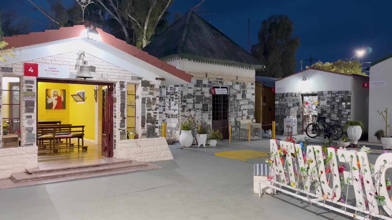 Escena de gran angular del santuario de la Difunta Correa al anochecer en San Juan, Argentina, con edificios de piedra iluminados, ofrendas florales, bancos y un ambiente crepuscular sereno.