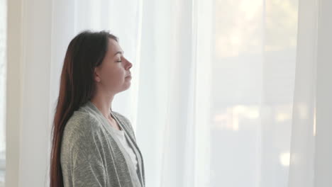 mujer con los ojos cerrados respirando profundamente mientras está de pie cerca de la ventana en casa por la mañana 1