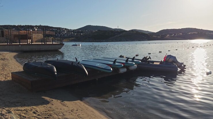 Motoscafi gonfiabili e kayak ormeggiati su una sponda soleggiata del lago con montagne sullo sfondo e acqua blu limpida.