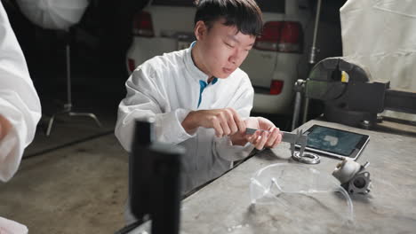 partial view of lady working with a microscope while young technician adjusts vernier caliper and taps on tablet in automotive workshop, featuring a carburetor on table and a white car parked