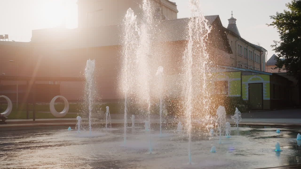 Amplia plaza con fuente a la hora dorada con destellos de sol, estanque reflectante y edificios de fondo, visitantes paseando cerca de los chorros y charcos, ambiente relajado de tarde de fin de semana y luz cinematográfica.