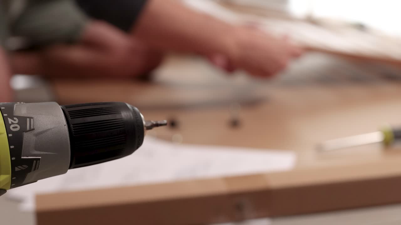 Hands assembling furniture using a power drill on a table. Soft lighting, blurred background, focus on tools and hands