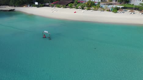 Lonely-Woman-on-Swing-in-Shallow-Water-in-Front-of-White-Sand-Beach-at-Maldives-Gulhi-Island,-Exotic-Tropical-Vacation-Destination,-Drone-Shot